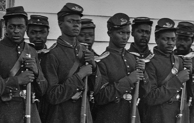 Eight United States Colored Troops, stand in two rows, with rifle barrels in their hands at their chests. One soldier is wearing a wedding ring on his left ring finger.
