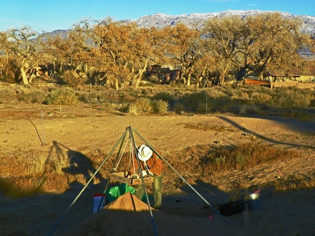 An archeologist sifts through the sandy soil of a test pit using a screen mounted on a metal tripod. In the background, snow capped mountains extend above trees.