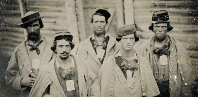 Four uniformed Confederate soldiers stare directly at the camera in this sepia photo. Each wears a small identification card suspended from a cord around their neck.