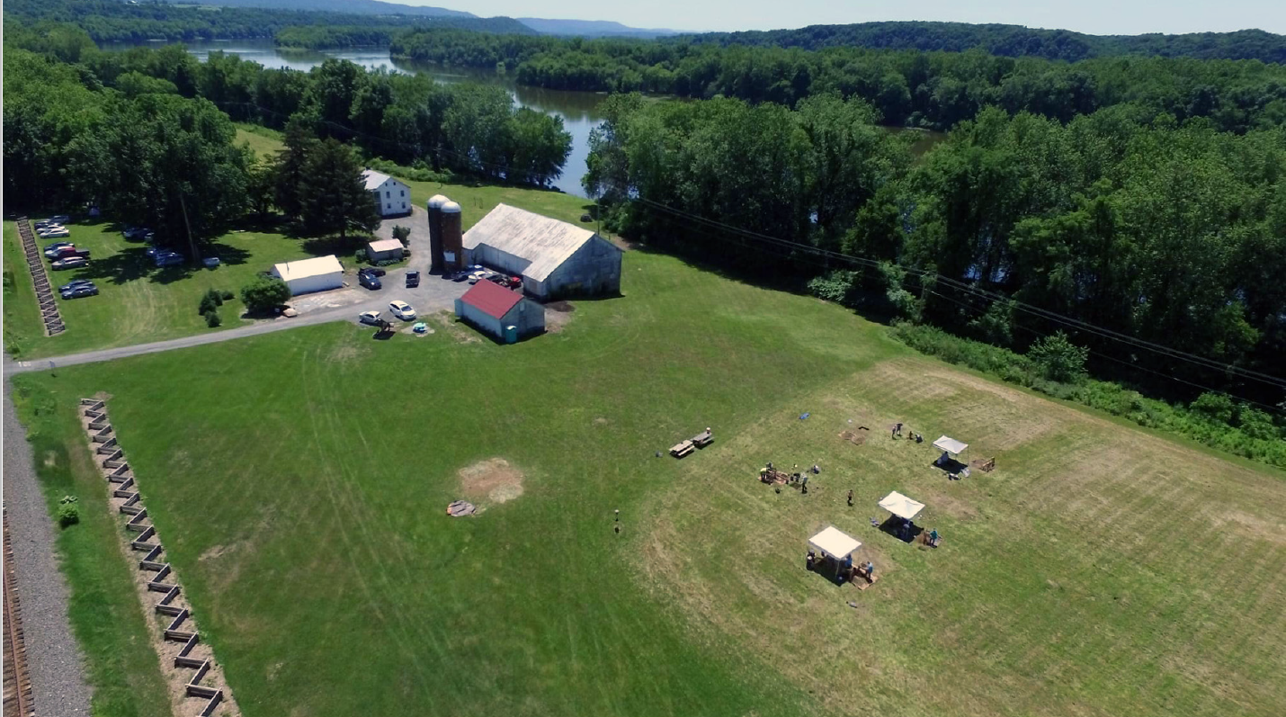 Three tents protect archeological test pits in a green field that lies between the banks of a river and a fence along railroad tracks with a farm in the background.