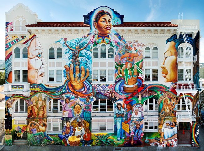 Facade of four-story, symmetrical white stucco building covered with vibrant mural celebrating women from various cultural and ethnic groups and across time