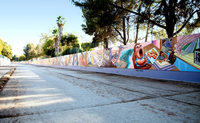 Angled view of long colorful mural on a low wall. The mural features representations of historical events from the 1940s. Concrete roadway visible in the foreground. Background features trees and a chain link fence.
