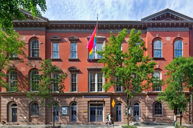 Three-story red brick building with brownstone elements. The facade is symmetrical and divided in three sections. There is a gay pride flag flying from the central window on the third floor.