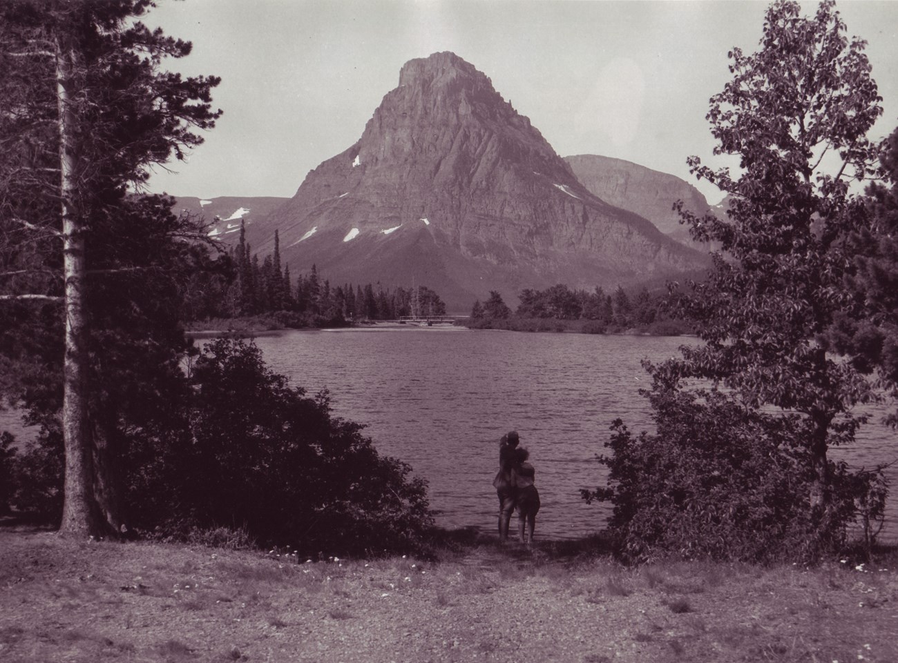 Black and white photo of a couple on the edge of a lake looking towards a mountain.