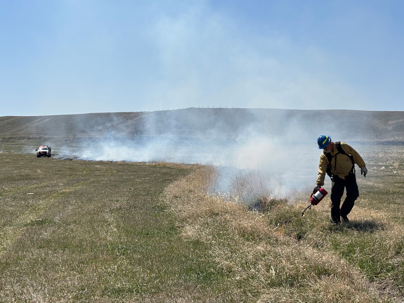 A firefighter uses a drip torch to remove grasses along ditches at Grant-Kohrs National Historic Site A wildland firefighter dressed in a yellow shirt, green pants, and a blue helmet lights grasses growing along a ditch in a grassy field. Smoke is rising from the ditch behind the firefighter; A fire engine with lights on is in the background.