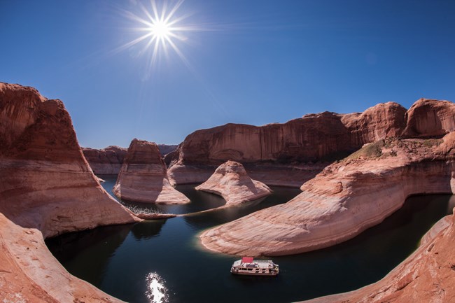 Dark green body of water winding through red rock formations with brilliant sun overhead