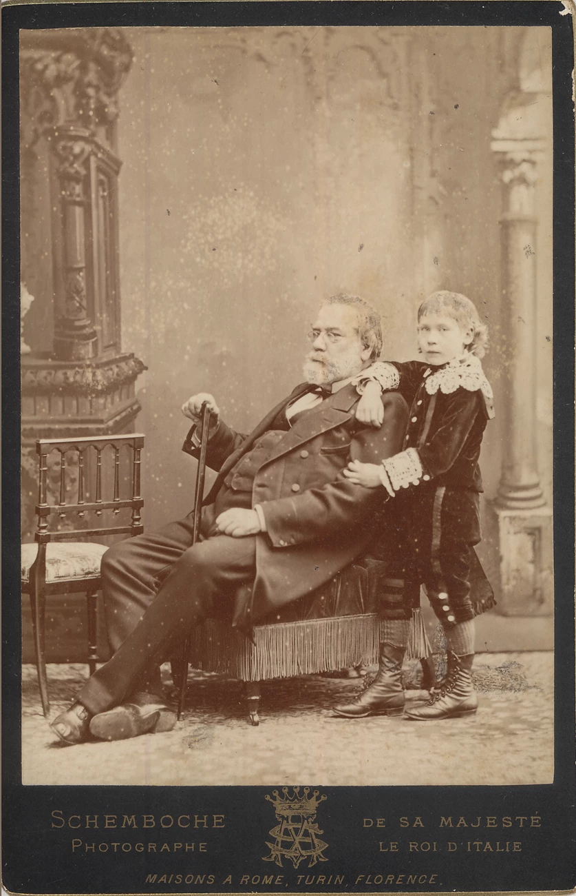 GPM with Carl and cane, c. 1875 historic black and white photo of older man sitting in a chair and a young boy standing behind him, text at bottom of photo reads "Schemborche Photographe, De Sa Majeste Le Roi D'Italie, Maisons A Rome, Turin, Florence"