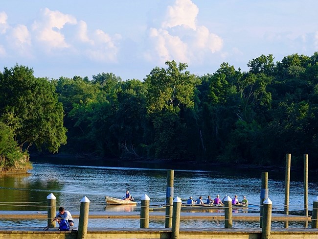A river landscape is surrounded by a forested edge and a blue sky with large, white clouds. A dock is in the foreground, with a man fishing off it. Behind the dock, people sit in a large rowboat.