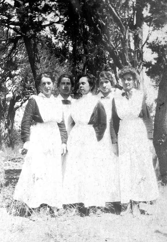 In an historic black and white photo, 5 Harvey Girl waitresses are posing outdoors for a group photo; their hair is pulled up and they are wearing black, long sleeve shirts with white full-length aprons.