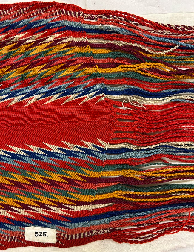 GRPO 525 The end of a woven sash with a zigzag pattern and braided ends. The dominant color is red with blue, green, brown, yellow, white zigzag stripes.