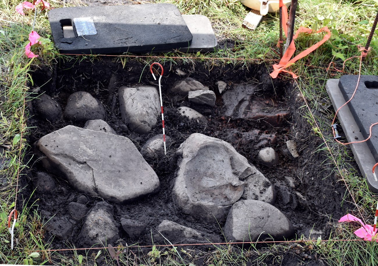 A square or earth with rocks showing. Around the edge are grass, orange flagging, and knee rests.