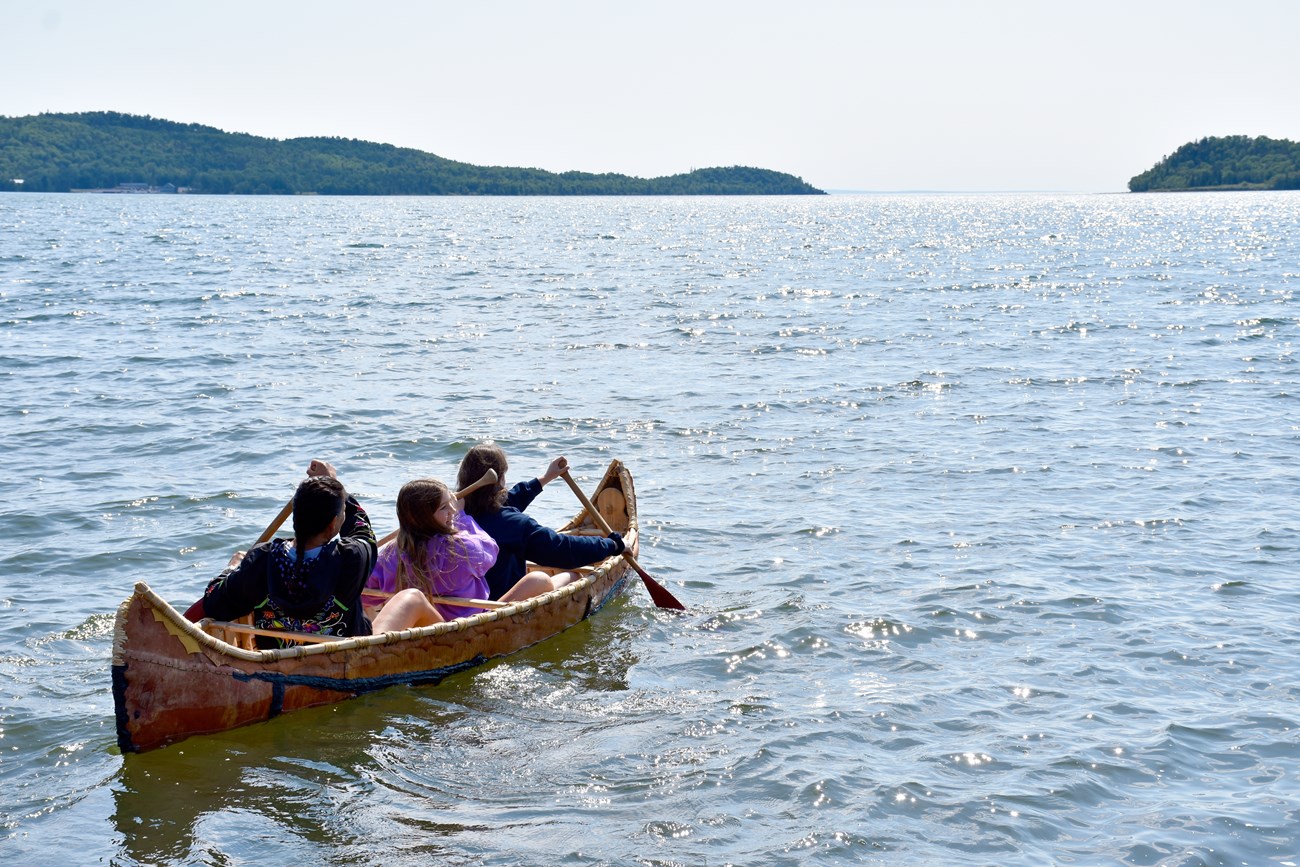 in the bay Three people paddle a birch canoe in a bay with distant landforms.