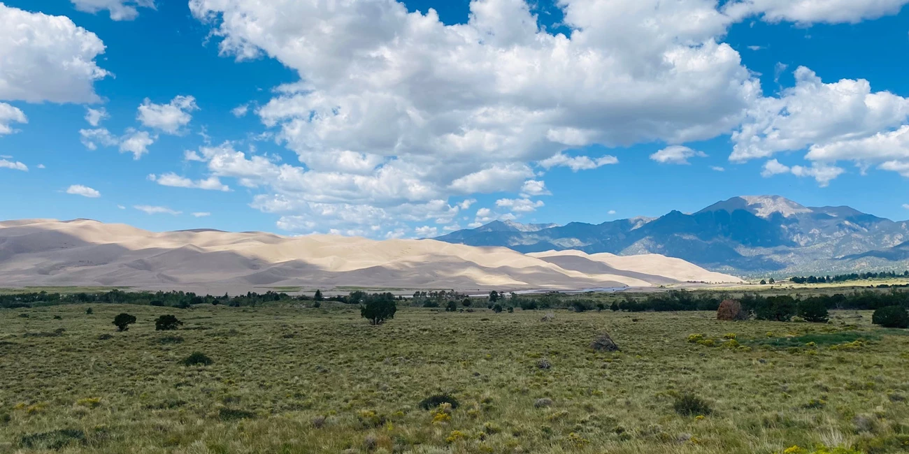 Great Sand Dunes In the mid distance, large undulating hills of sand appear like small mountains backed up against taller granite mountains flecked with green aspen and pine forests.