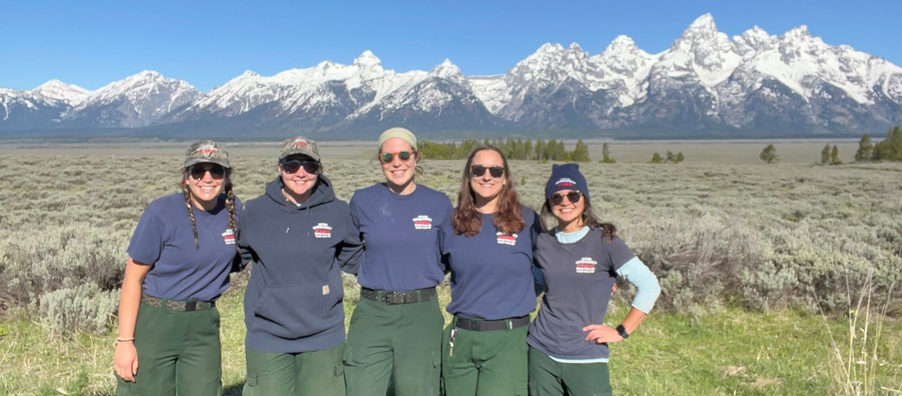 Participants at the academy at Grand Teton National Park. Five people in crew shirts smile in front of the Tetons.