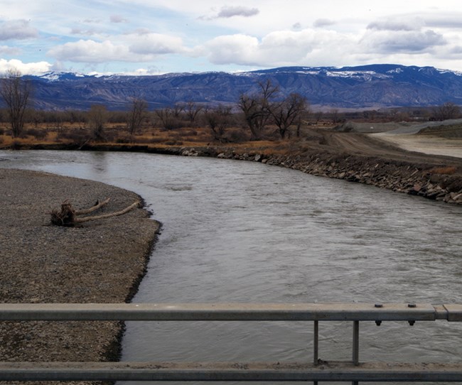 A river with rock and dirt banks viewed from a bridge with mountains in the distance.