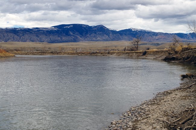 A bend in a river lined by rocky shores and dirt banks with snow tipped mountains in the distance.