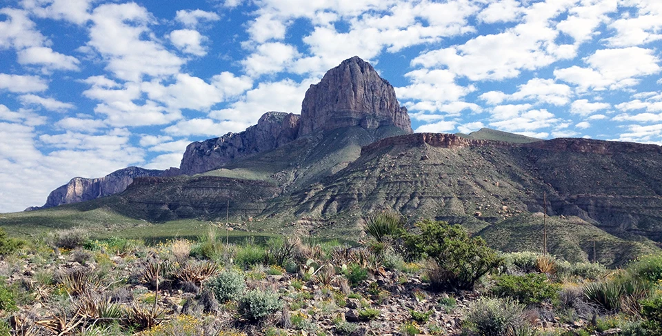 Guadalupe Mountains National Park Mountain peaks of sheer rock faces above a shrubby desert floor under scattered, cottony clouds.