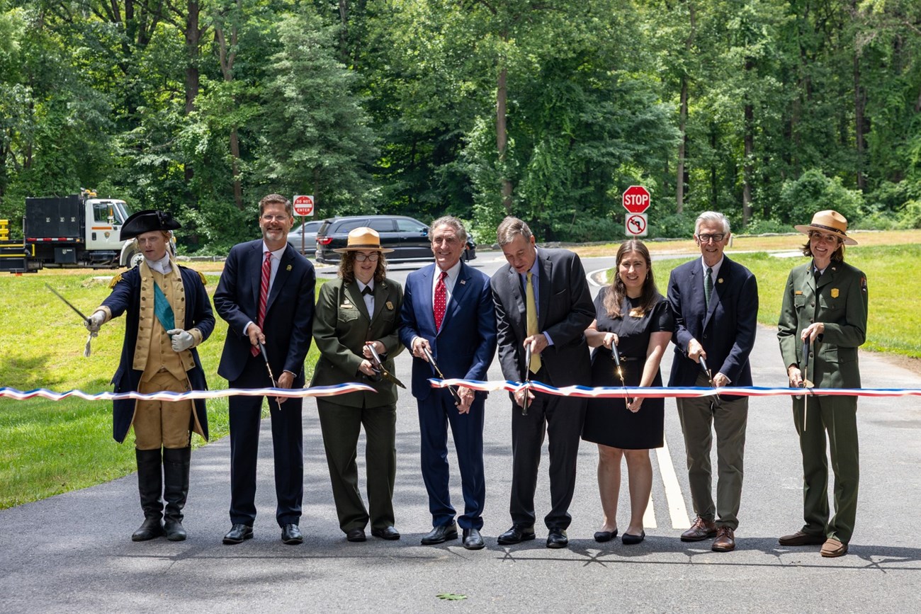 Several dignitaries cut a red white and blue ribbon on a roadway.
