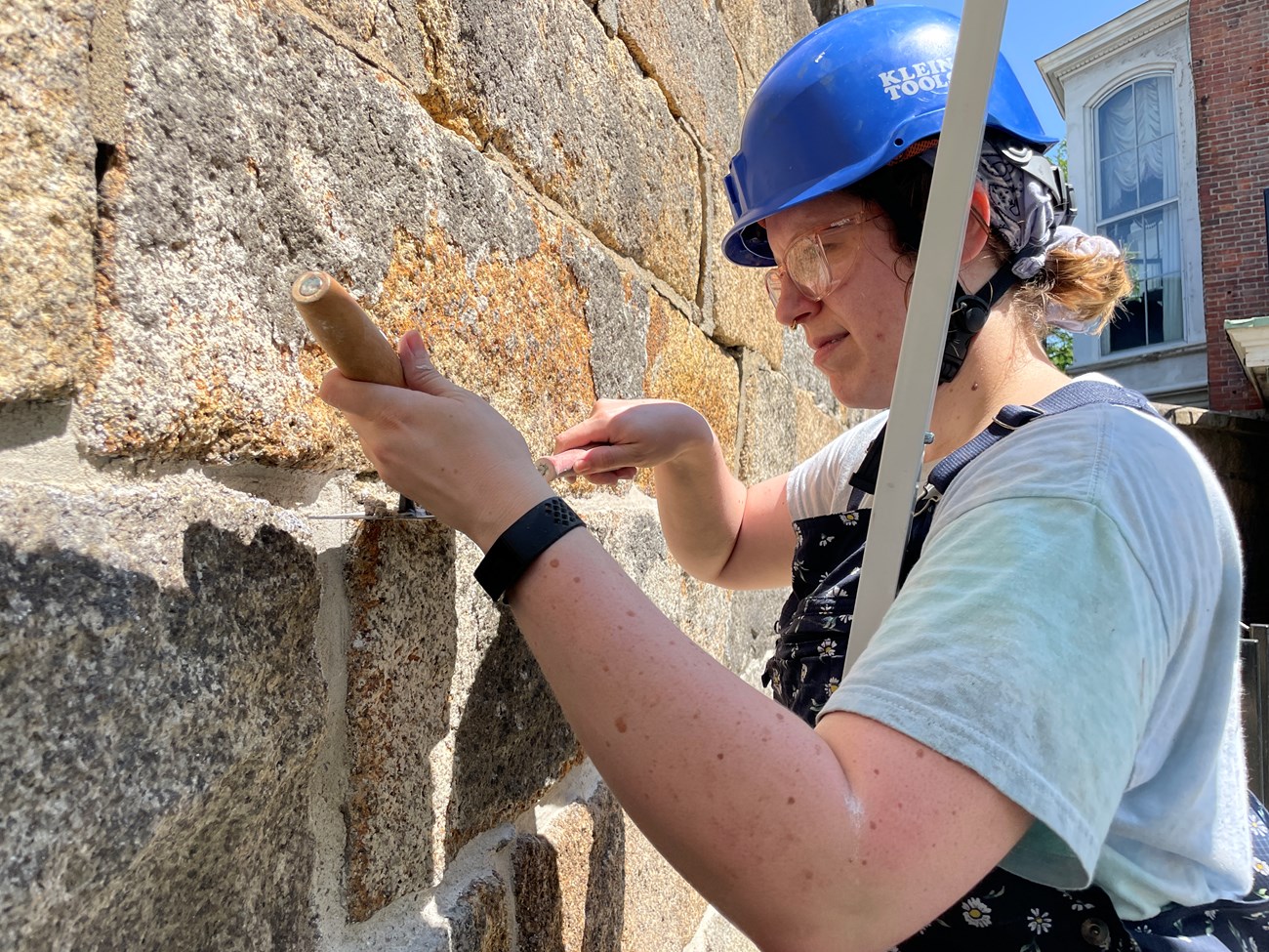 A closeup of a crew member wearing a hard hat standing close to a granite block wall. They are holding a masonry trowel with mortar in one hand and a masonry jointer in the other and pushing mortar into an empty space between the blocks.