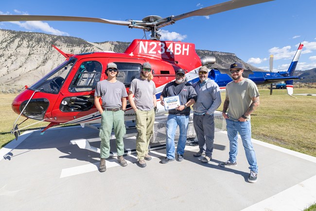 A group of men, middle one holding plaque, stand in front of red, white, and blue helicopter near a mesa.