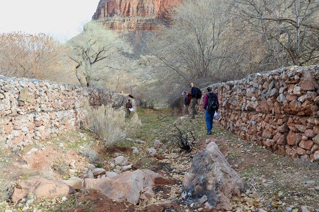 Four people stand in between two parallel gabion walls constructed with rocks.