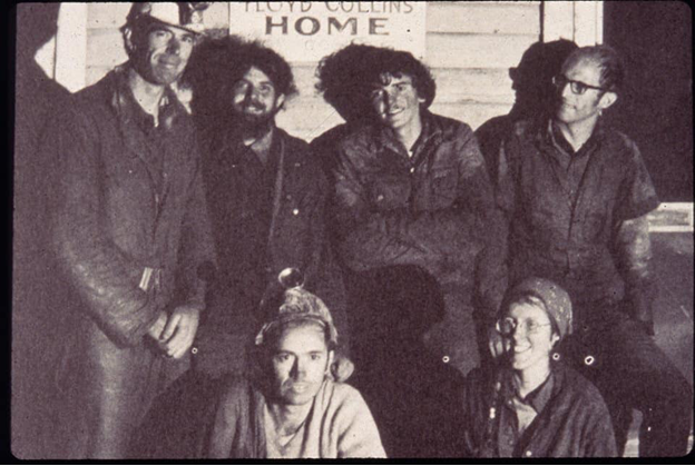 A black and white photo of six cavers posing for a group photo. They are wearing caving gear covered in mud.