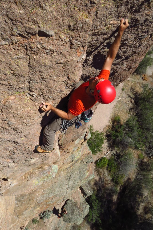 View from above of Gavin in a red helmet and climbing shoes, scaling a tall rock formation. A rope trails dozens of feet below him towards a tiny-seeming person on the ground.