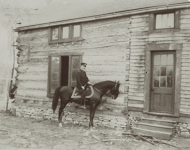 Black and white photo of a man on a horse in front of a log cabin