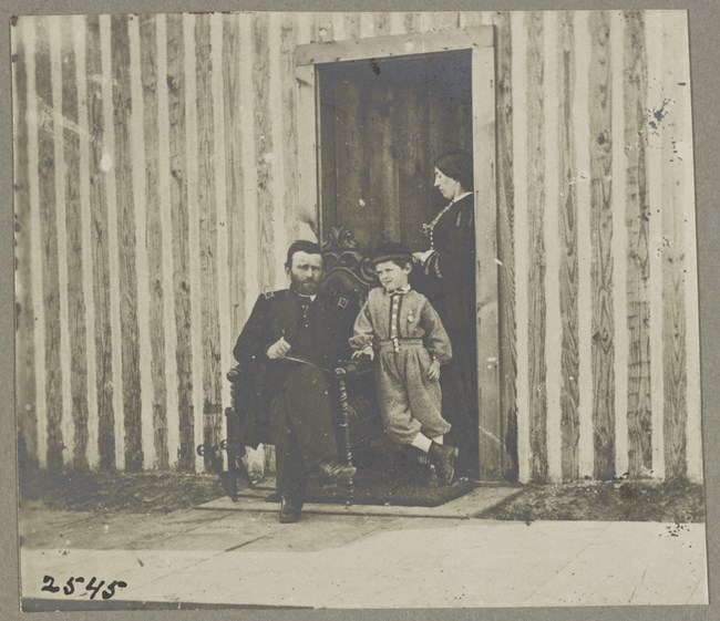 Black and white photo of a man in uniform sitting on a chair with a young boy leaning on him in front of a doorway where a woman is standing in side profile