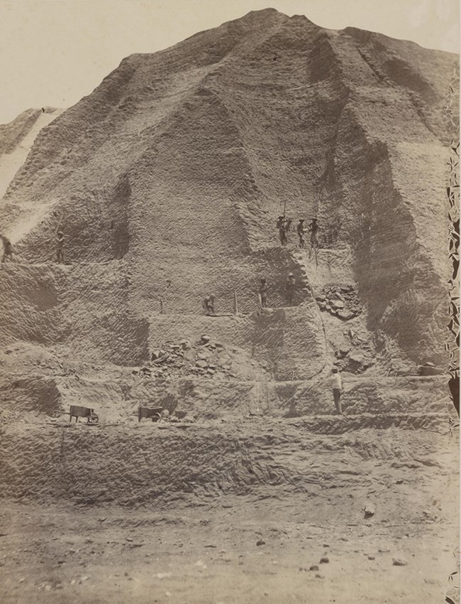 Black and white photo of a large mountain of guano being mined by Chinese workers. Several workers are dwarfed by the size of the mountain of guano. Wooden wheelbarrows wait at the base to transport the guano.