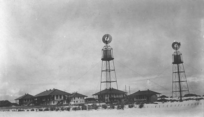 Black and white photo of the cable station, including four 2-story buildings with dark roofs, and what looks like a bungalow. Two towers with what look like water tanks and windmills are in the foreground. Low scrub and sand surround the buildings.