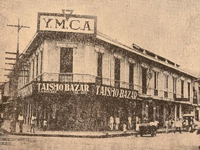 Black and white photo from a newspaper. Taken from the street, there are several people and carts in front of the Taisho Bazar building. The second floor has a large YMCA sign.