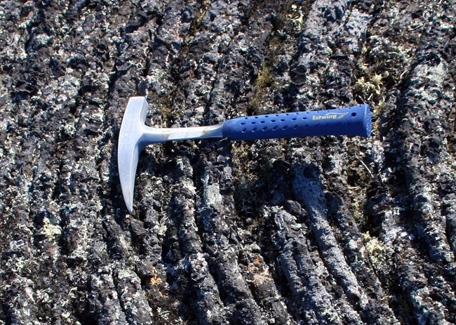 Silver hammer with sharp, arched, pick-shaped head and a blue handle sits on volcanic rock.