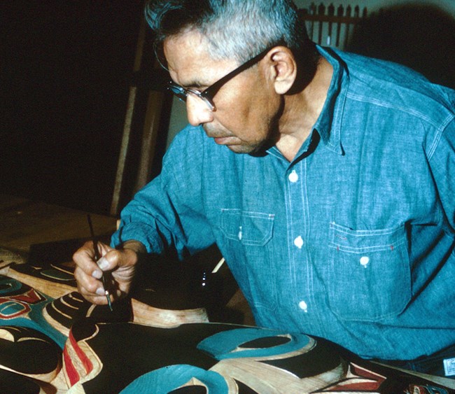 A man in a blue shirt holding a paintbrush. A totem pole in laying in front of him.