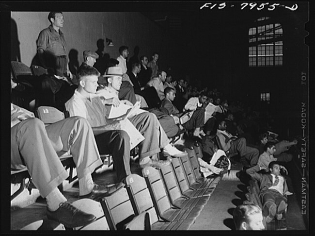 Black and white photo of young white and Black men sitting in tiered auditorium seating. Some of them are waiting attentively while others lounge in their seats. One man at right sits sideways and stretches his legs across multiple seats.