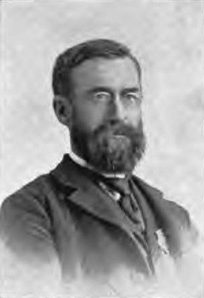 Portrait of man in suit and tie. Dark hair, trimmed beard and mustache, small wire glasses.