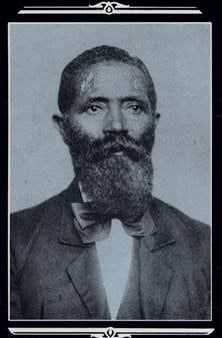 George Teamoh Black and White photo showing a man from the waist up staring to the right with his mouth in a neutral line. He is wearing a suit jacket, bow tie, and vest.
