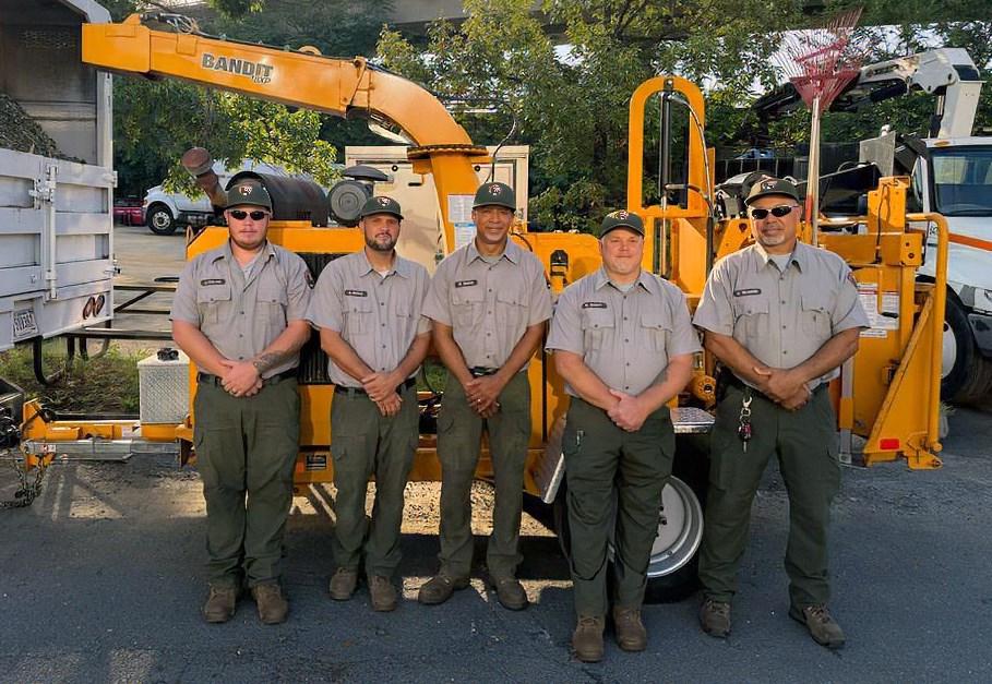 Five men in grey and green uniforms and green ball caps with hands folded in front of a yellow trailer with rakes attached to a white truck filled with green plant material