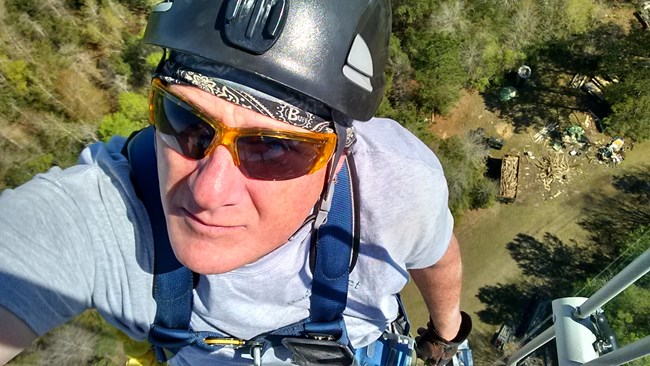 Tim Gilk, Big Thicket Tower (300-feet) A man wearing a helmet, sunglasses, and climbing gear takes a selfie from the top of a 300 foot tower.
