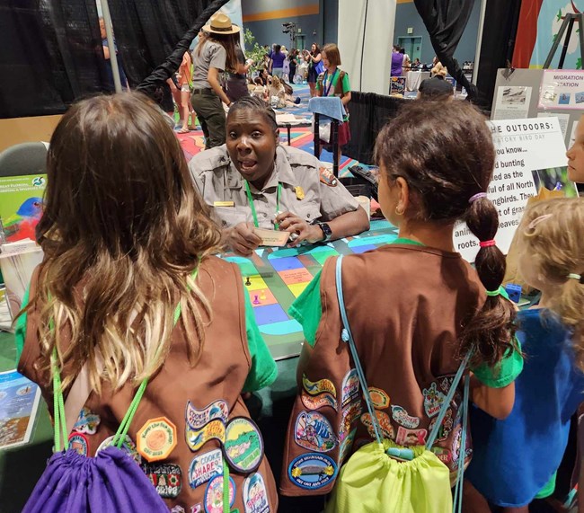 A Black woman sitting at a table in uniform explaining a game to three young girls