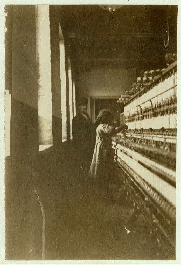 Girl in mill Yellowed photo of a young girl at work at a textile mill machine as man in a suit and hat looks on.