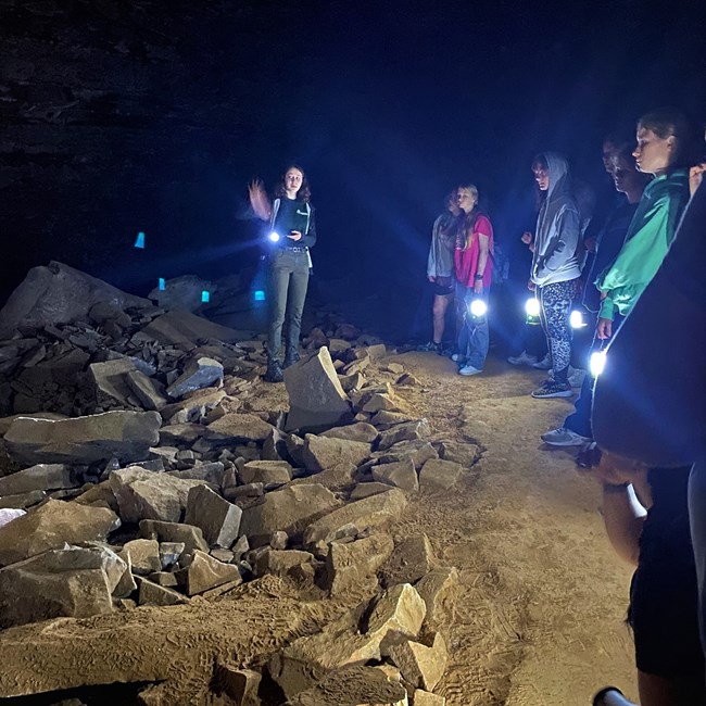 Campers explored the history of cave tour routes that have been in use since the early 1800s with Mammoth Cave’s Greening Youth Intern Alena. A person talks to a group of young women holding lanterns inside a rocky and dark cave.