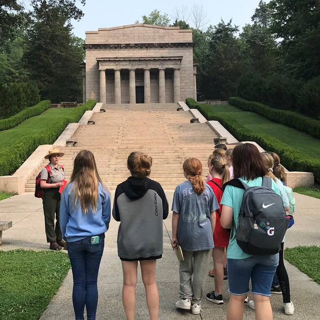 Following Their Footsteps campers explore Abraham Lincoln Birthplace National Historic Park with Ranger Noelle and visit the Memorial Building which protects the Symbolic Birth Cabin of Abraham Lincoln. A group of people face a long and wide stairway that leads to a large stone building with large columns.