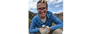 Kimber in a blue shirt and glasses, holding a falcon nestling with both hands.