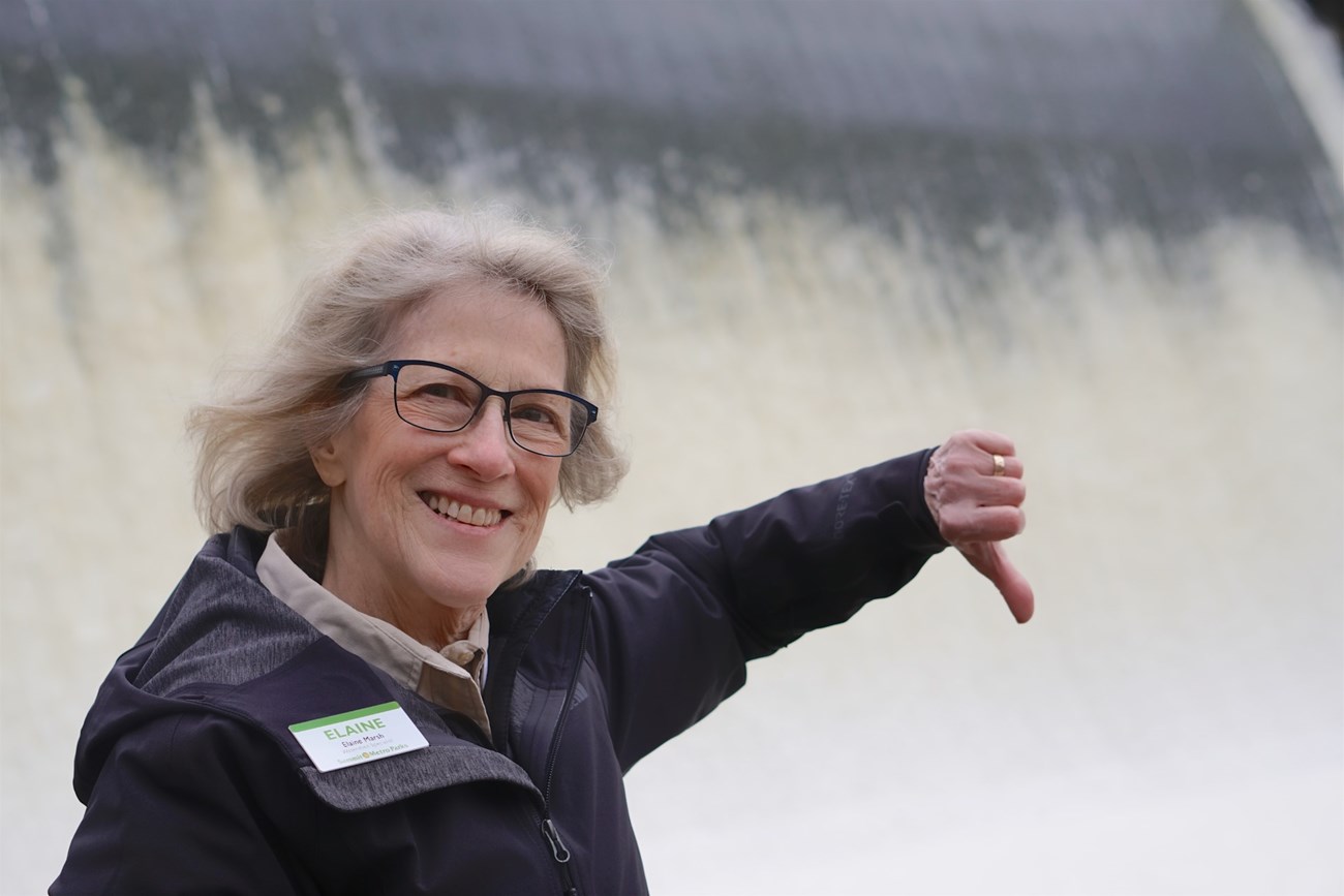 A woman smiles and gives a thumbs down to the wall of rushing water behind her. She wears glasses and a Summit Metro Parks nametag.
