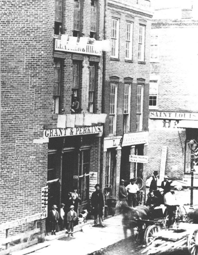 Black and white photo of brick storefront and street with horses and people in 19th century clothing