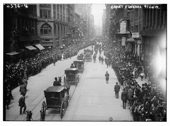 Black and white photo of the back view of a funeral procession of horses and carriages going up a street with crowds along the sidewalk watching