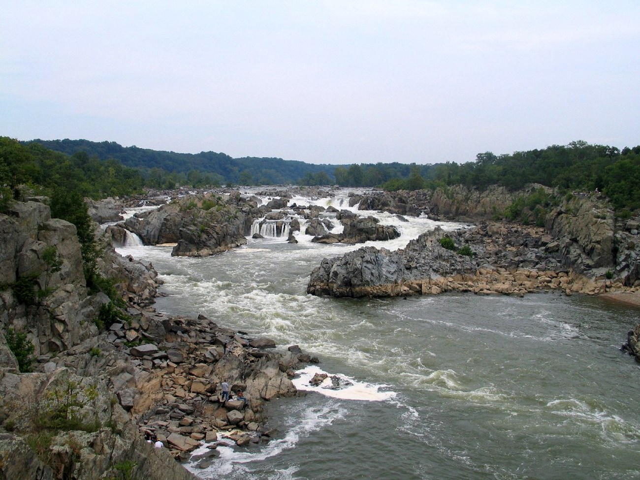 A river winds through a rocky landscape. The rocks are gray and beige in color. A forest and blue skies are in the background
