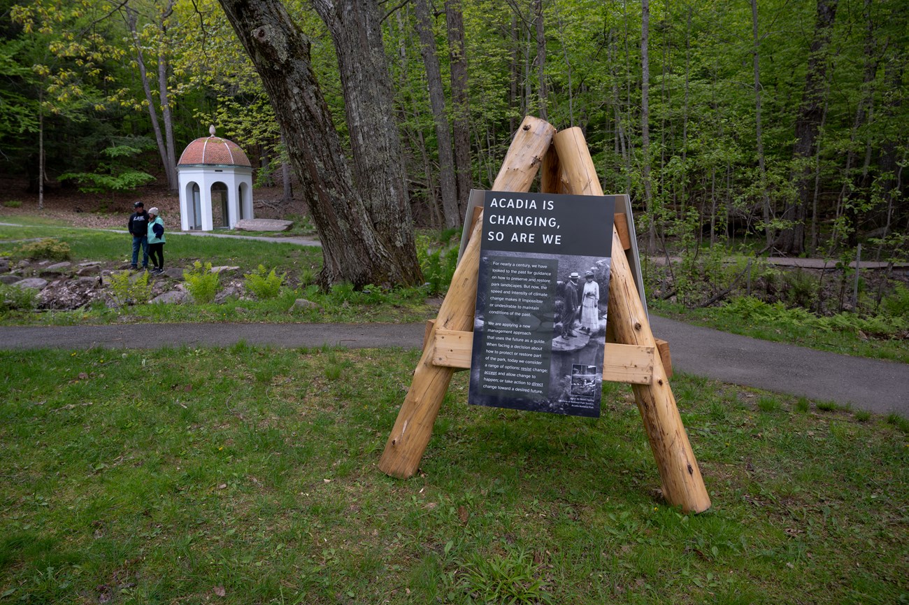 Wood structure with three legs and display panels attached is placed on a lawn next to a pathway with a historic domed structure and two visitors standing in the distance. Wood structure with three legs and display panels attached is placed on a lawn next to a pathway with a historic domed structure and two visitors standing in the distance.