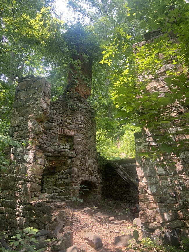 The ruins of the two-story home built by the Green family. It consists of portions of cobblestone walls with a large rectangular red brick chimney. Green foliage surrounds the ruins.